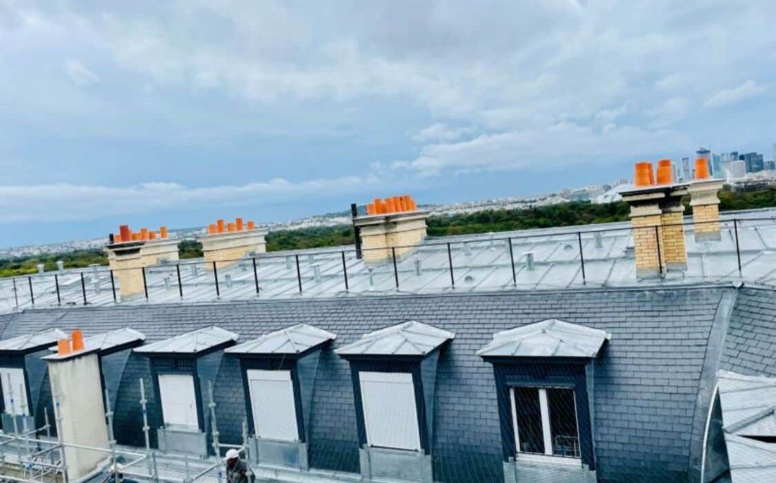 Toits en ardoise avec cheminées et ouvrier, ville en fond.